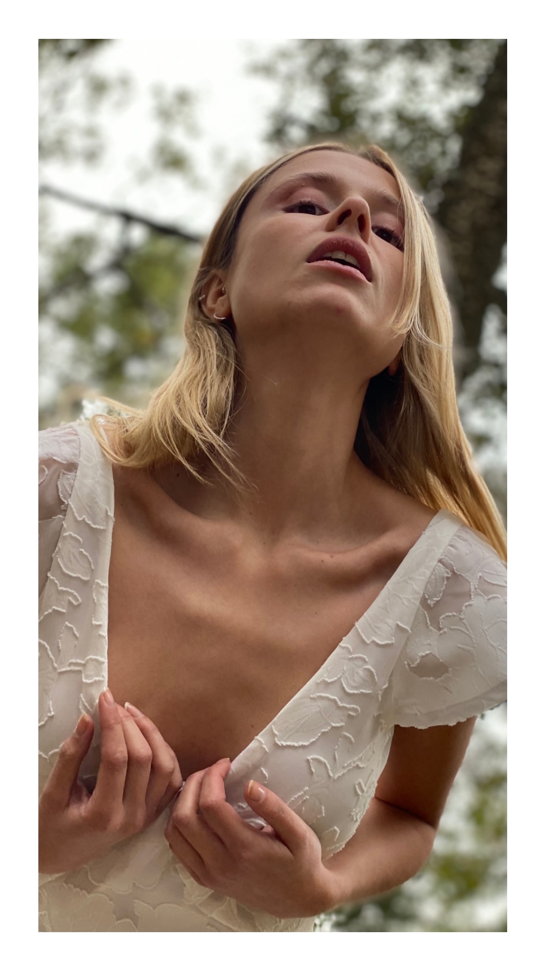 Low-angle outdoor portrait of a blonde woman in a white embroidered dress holding its neckline.