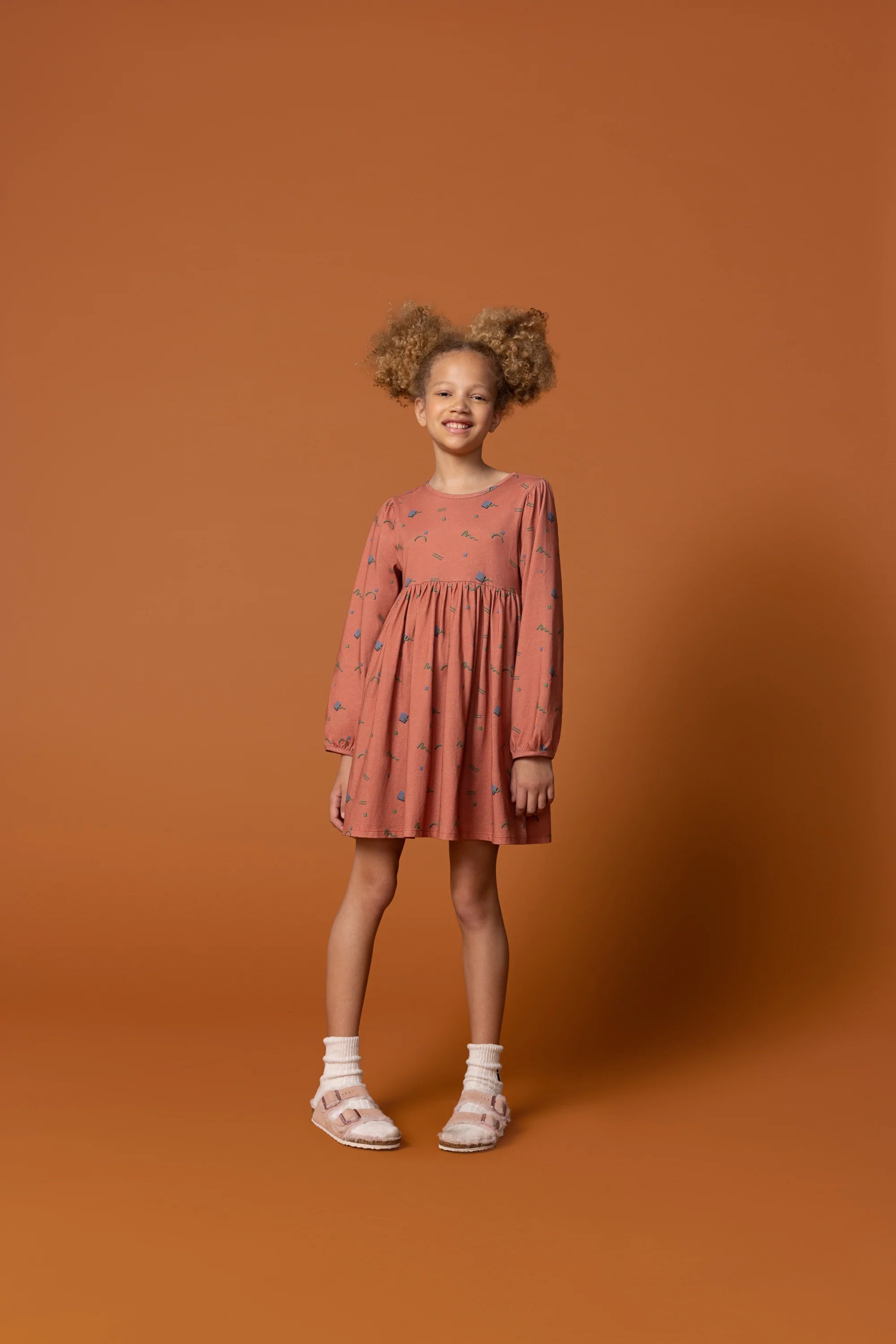 Smiling girl in a patterned coral dress, white socks, and strapped shoes against an orange studio background.