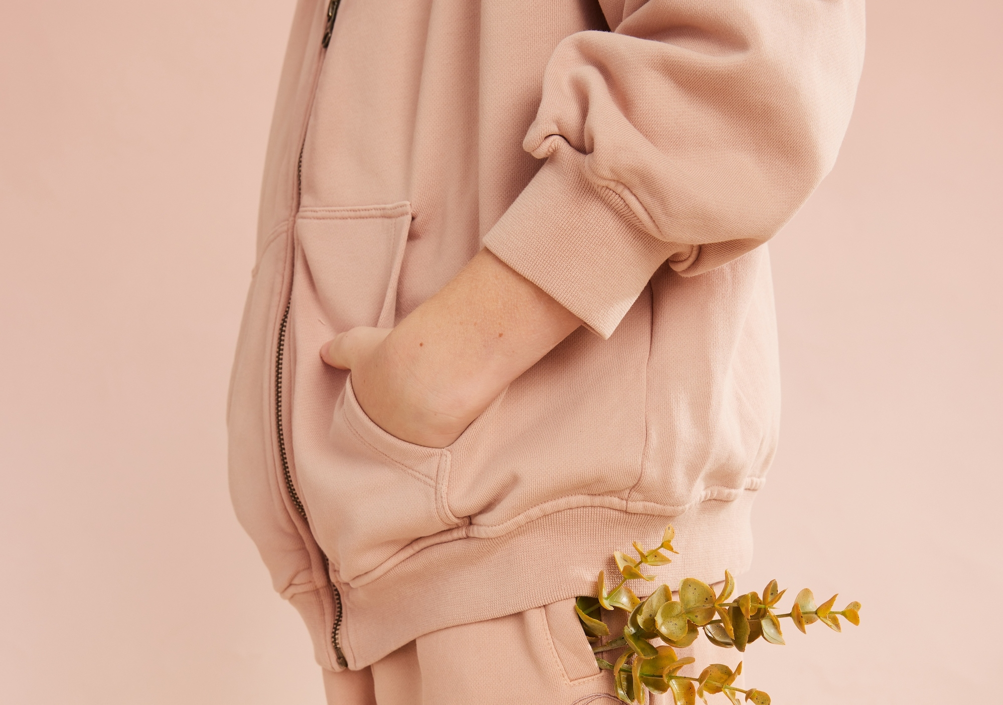 Cropped person in a blush-pink zip jacket with a hand in its pocket and a leafy plant at the waist.
