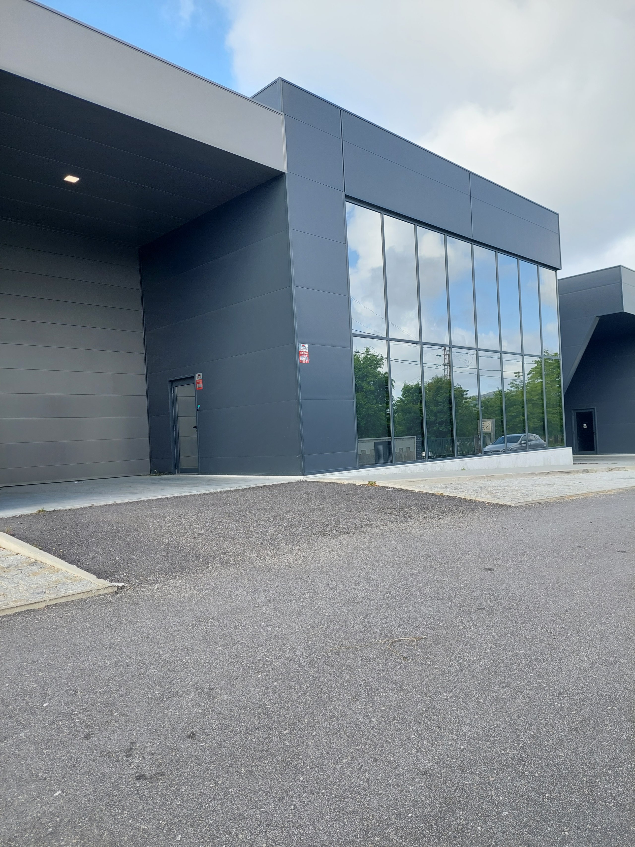 Modern dark gray commercial building with reflective windows, security stickers, asphalt foreground, and cloudy blue sky.