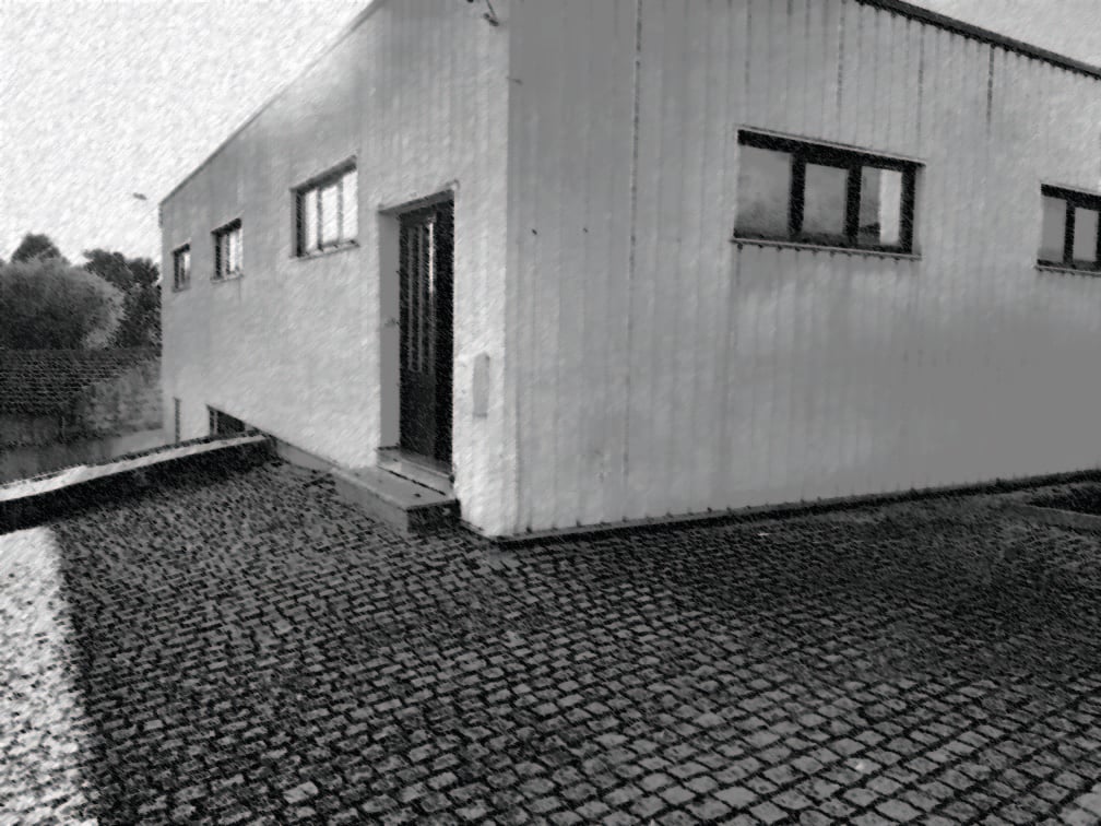 Blue-green photograph of a plain building with dark windows and doorway beside a cobblestone courtyard.