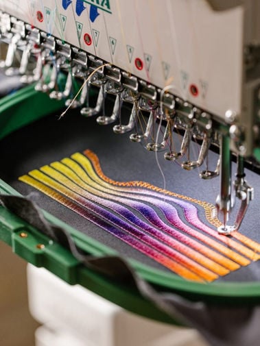 Multi-needle embroidery machine stitching colorful wavy bands into fabric held in a green hoop.