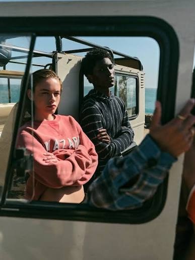 Two passengers sit inside an older vehicle by the sea while a plaid-sleeved person holds the window frame.