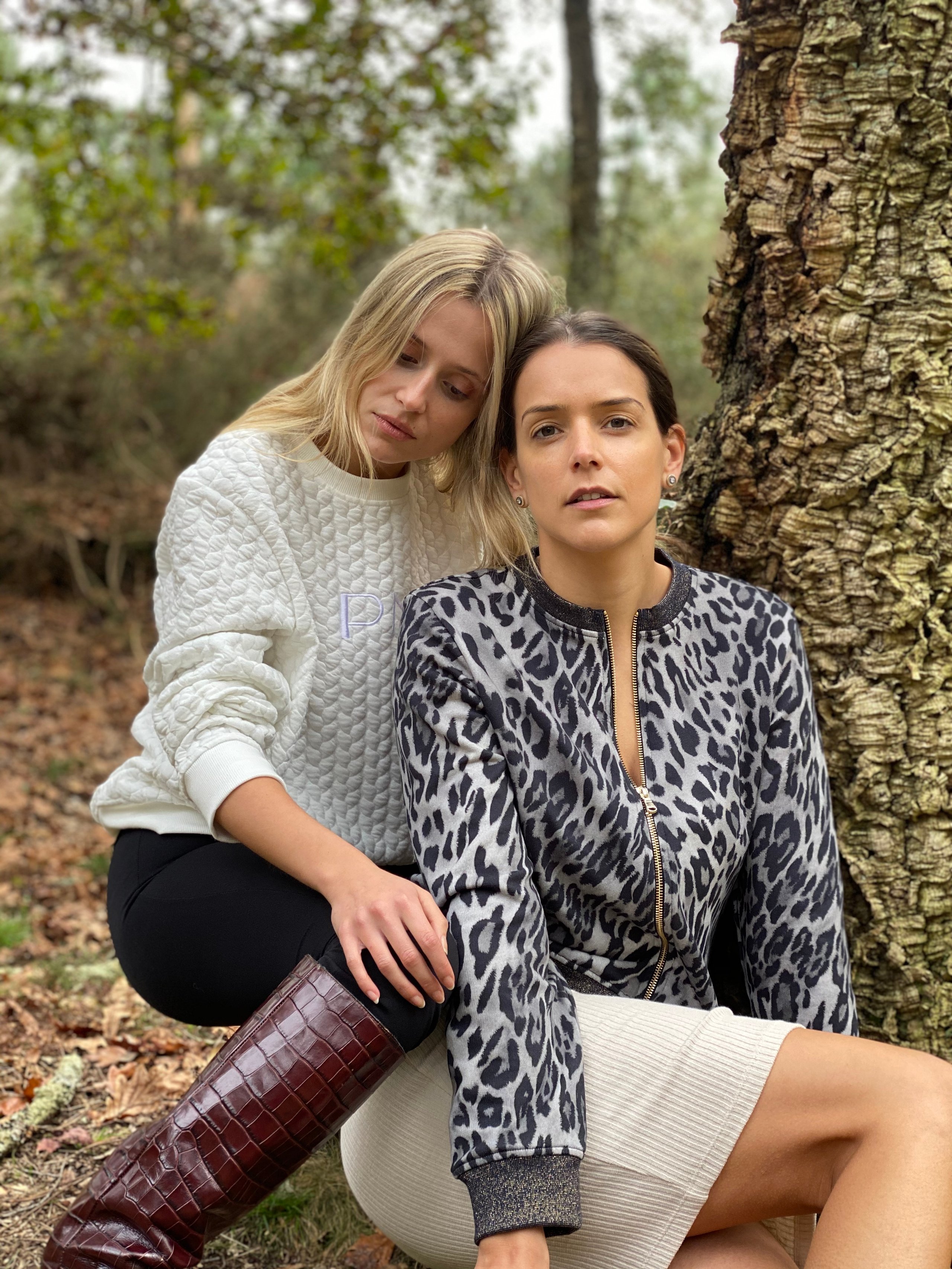 Two women pose beside a deeply textured tree trunk in a softly blurred forest.