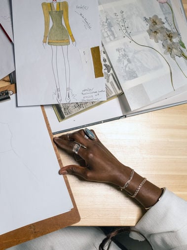 Top-down view of a fashion sketch, botanical reference book, blank drawing board, and artist’s hand on a wooden desk.