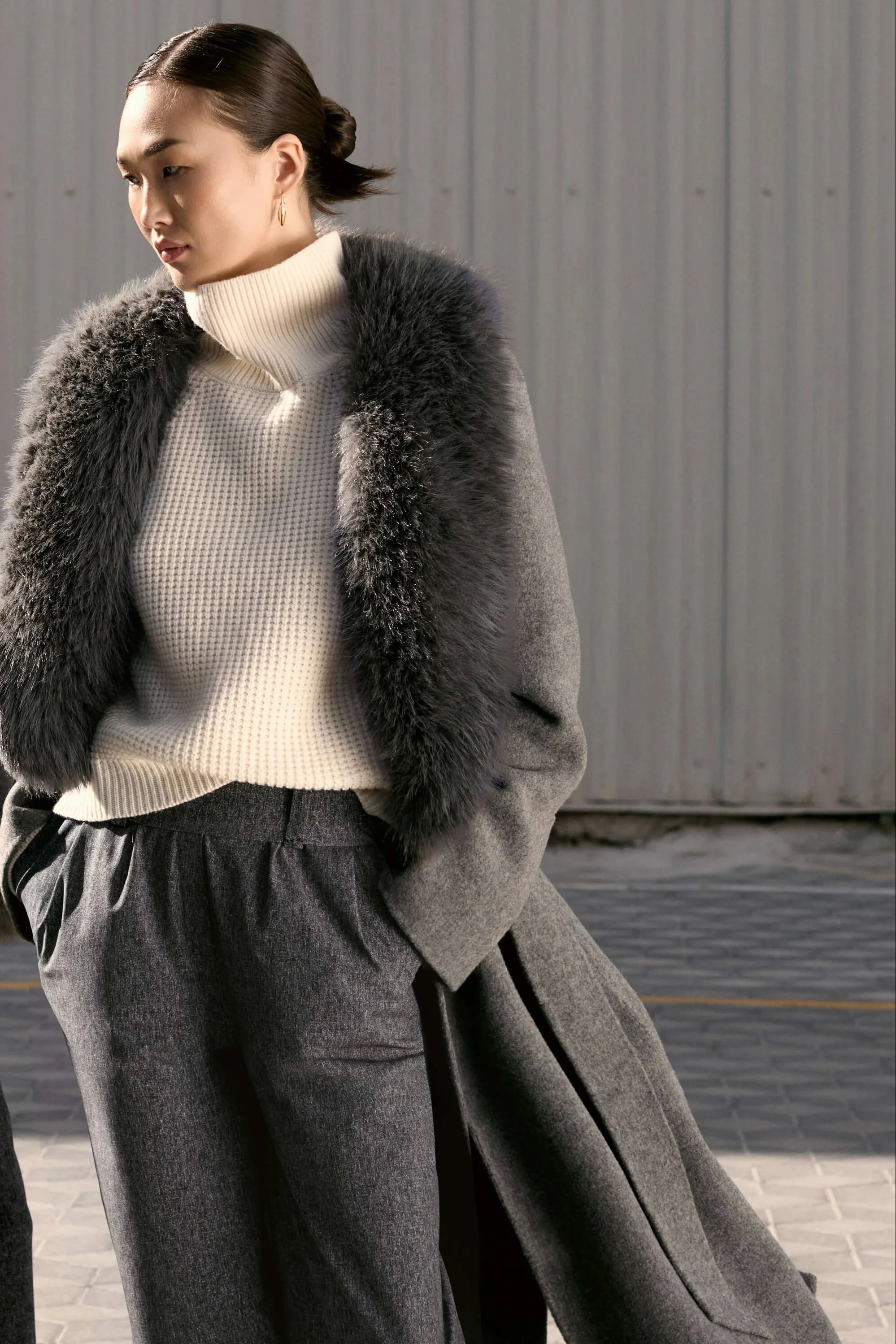 Woman in a cream turtleneck, gray coat with black fur panels, and pleated skirt against a gray wall.