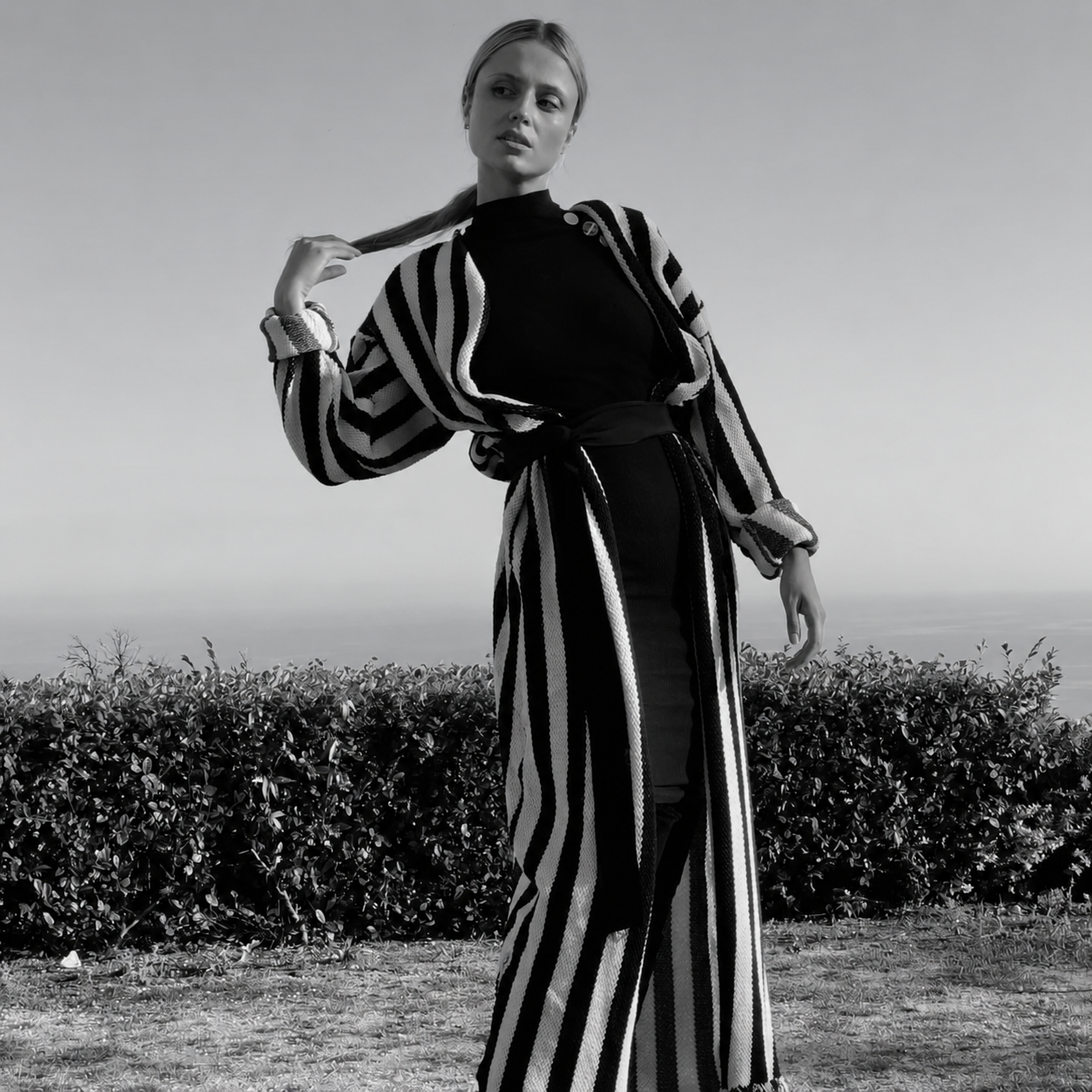 Woman in a striped long cardigan standing beside a hedge in a black-and-white outdoor fashion photograph.
