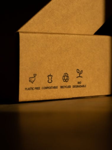Open brown cardboard box with four eco-label icons reading “PLASTIC FREE,” “COMPOSTABLE,” “RECYCLED,” and “BIO DEGRADABLE.”