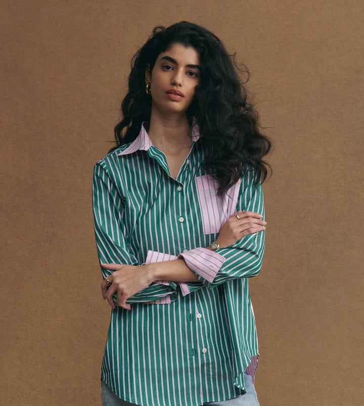 Woman with long dark hair wearing a green-and-white striped shirt with pink accents, standing against a brown backdrop.