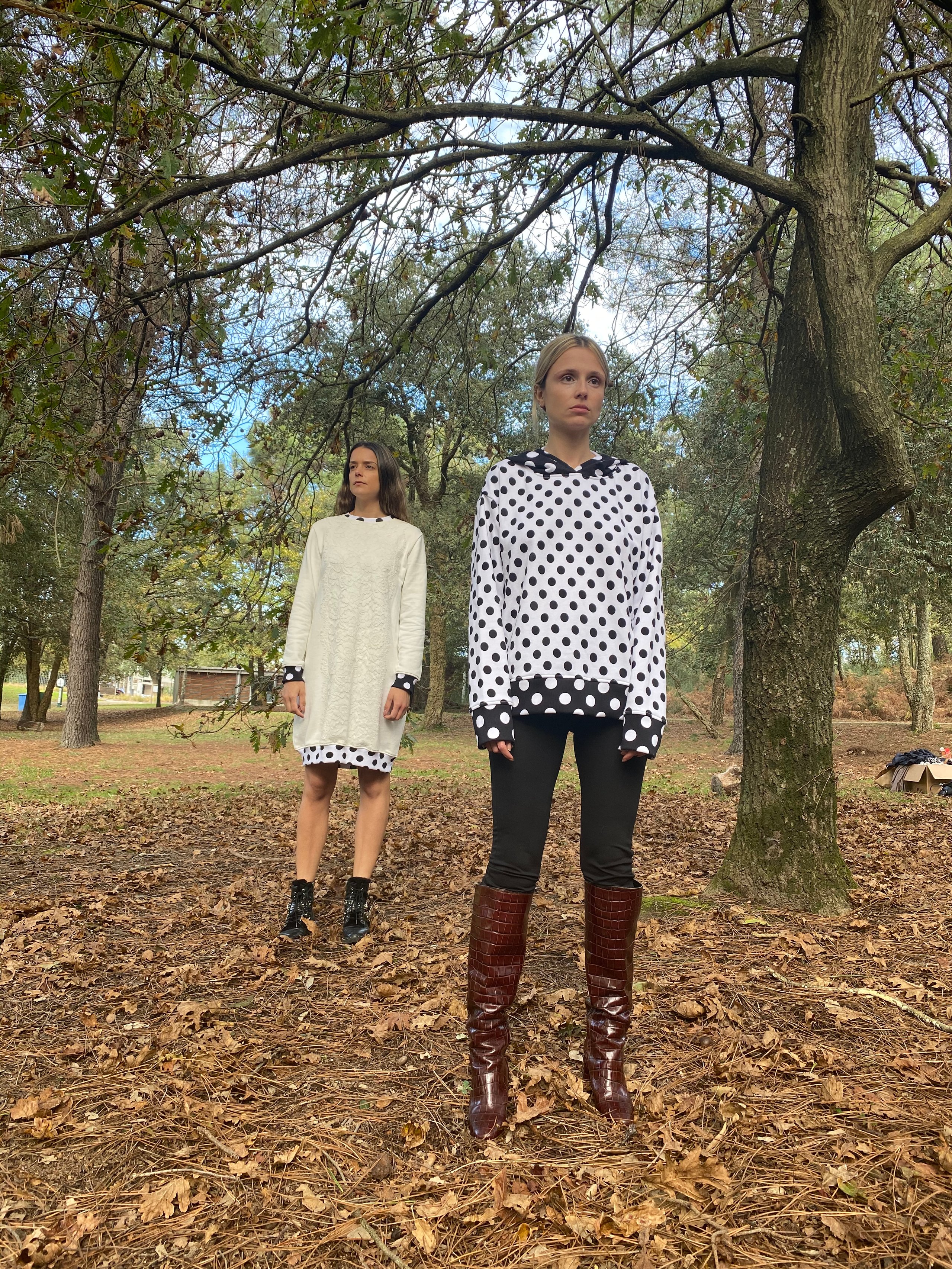 Two women in black-and-white patterned clothing stand beneath trees in a leaf-covered wooded area.