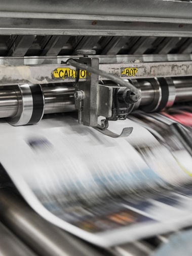 Close-up of metal printing press rollers feeding a blurred, multicolored printed sheet.