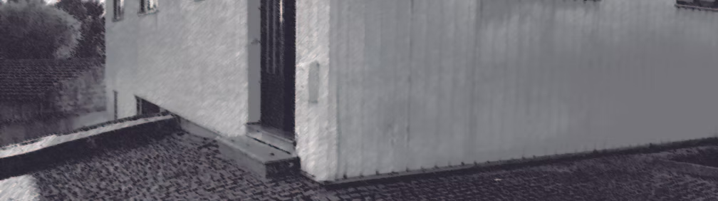 Grayscale exterior view of a pale building corner with a dark narrow opening, trees, and cobblestone paving.