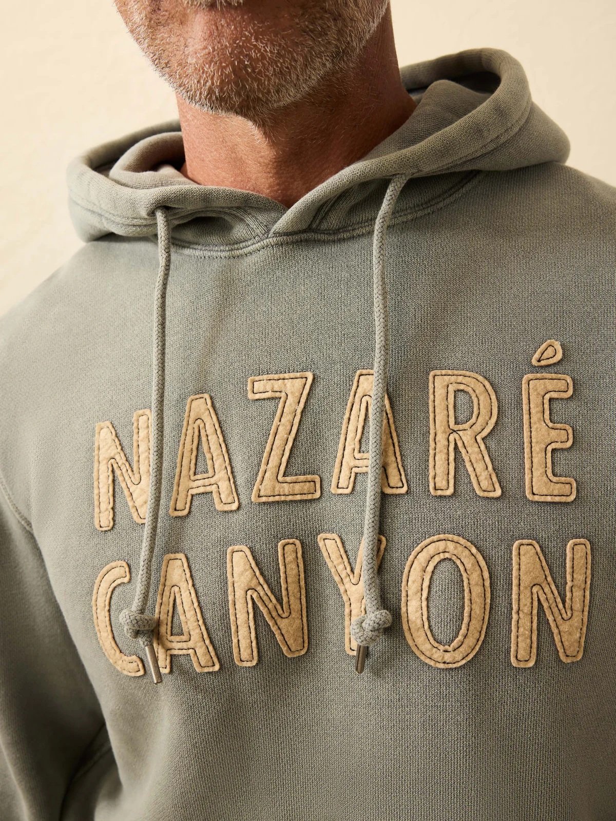 Close-up of a gray-green hoodie with tan stitched “NAZARE” and “CANYON” lettering.