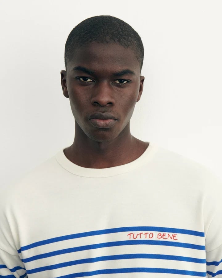 Front-facing portrait of a young man wearing a white shirt with blue stripes and red “TUTO BENE” text.